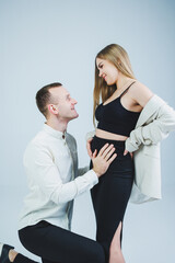 Smiling young man kneeling with his pregnant wife and hugging her belly, isolated on white background. Stylish young people. A man in a shirt and a woman in a black skirt and jacket