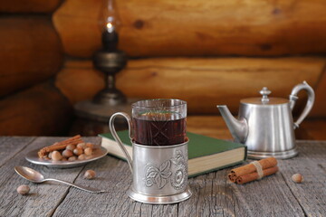 On wooden table on background of old lantern standing on wooden barrel near log wall is glass of tea in iron holder next to iron plate with nuts, iron teapot and book. (Text of book is in English.) 