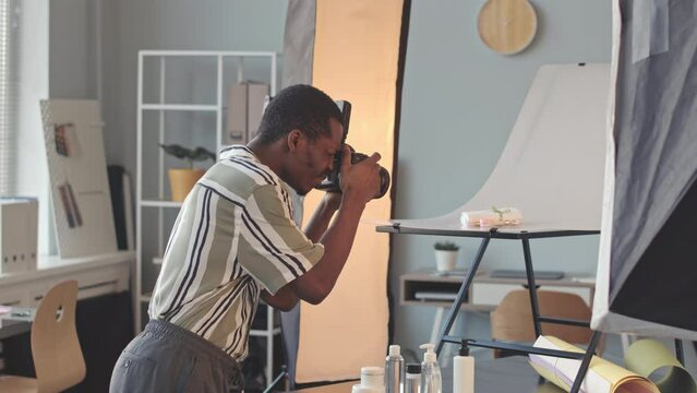 Medium slowmo of professional African American male photographer doing commercial photoshoot for cosmetic brand, taking pictures on digital camera of skincare products on white background