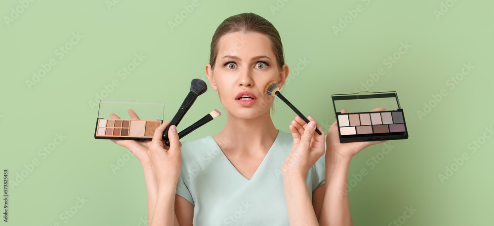 Sticker shocked young woman and hands with makeup brushes and decorative cosmetics on green background