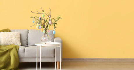 Vase with tree branches and Easter eggs on table near sofa in room interior. Banner for design