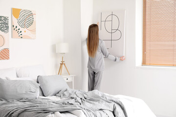 Young woman hanging poster on light wall in bedroom