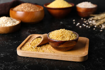 Wooden board with bowl and scoop of bulgur on dark background