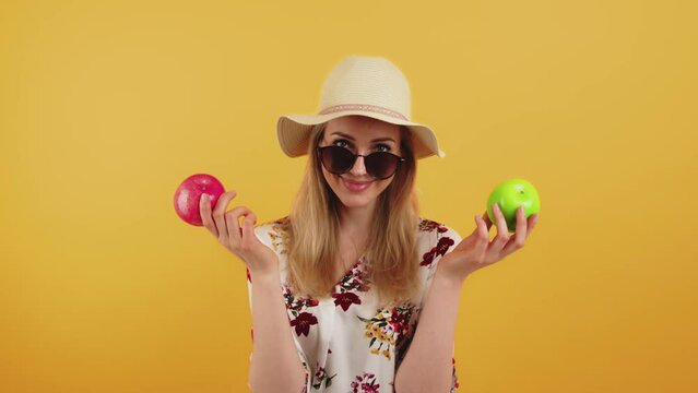 A smiling young woman wearing sunglasses and a hat holding red and green apples. High-quality 4k footage