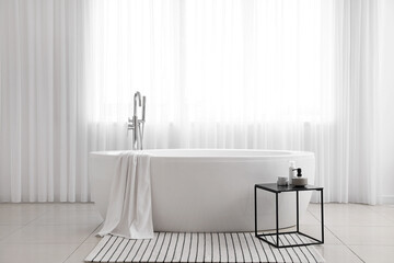 Bathtub and table with cosmetic products in light bathroom