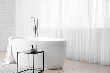 Bathtub and table with cosmetic products in light bathroom