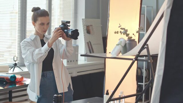 Medium slowmo of professional female still life photographer taking pictures on digital camera of minimalist composition, working in bright spacious photo studio with lots of photography equipment