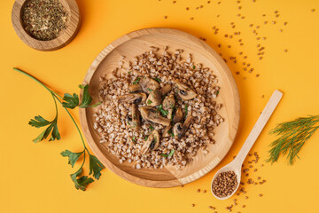 Wooden plate of tasty buckwheat porridge with mushrooms, parsley and dill on yellow background