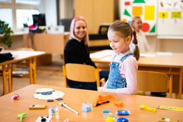 The teacher leans over the student. A girl sits in a school desk and the teacher checks her work.