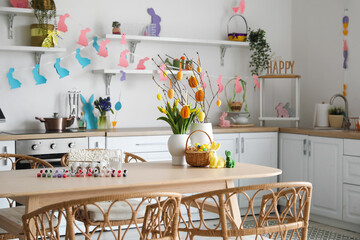 Vases with tree branches, Easter eggs, tulips and rabbits on dining table in kitchen