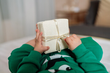 Baby gift. A little boy in a green suit holds a gift box with his feet and hands in bed.