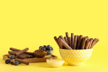 Bowls with delicious chocolate wafer rolls and condensed milk on yellow background