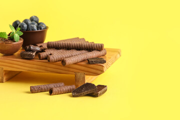 Naklejka premium Board with delicious wafer rolls, chocolate pieces and blueberries on yellow background