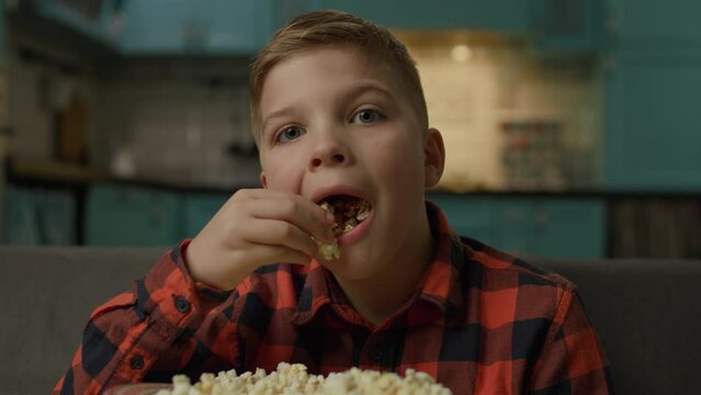 Child Eats Popcorn Looking At Camera. Boy watching TV with popcorn sitting on sofa POV. Kid eating close up.