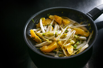 Fried chicory with orange pieces and spring onions in a black frying pan on the cooker, healthy...