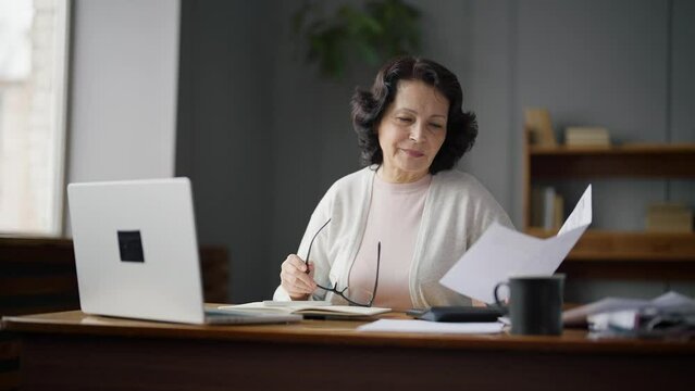 An elderly woman sits at a desk at home and studies payments or taxes. She checks the numbers in the documents and enters the data into the laptop. The concept of bill payments and tax calculation.