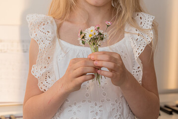 A blonde girl holds a bunch of daisies in her hands, she is dressed in a white dress, stands...
