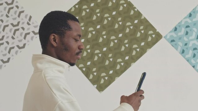 Young African American man visiting picture exhibition in modern art gallery taking photos on smartphone of unusual art objects and patterns