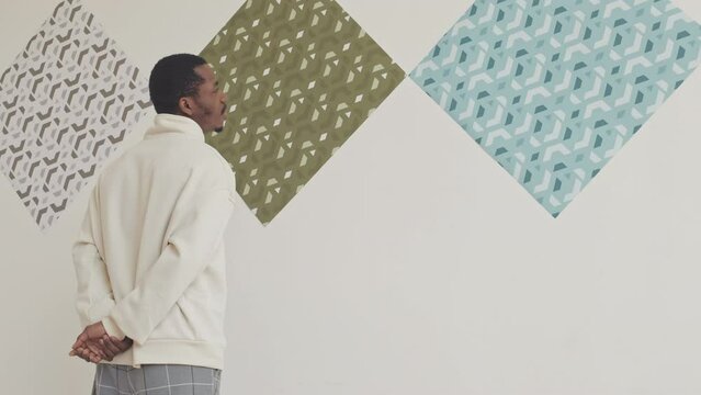Black man examining patterned abstract pictures hanging on minimalist white wall in modern art gallery