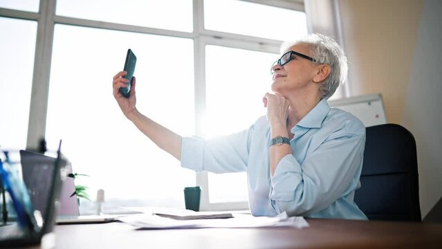 Happy businesswoman wearing eyeglasses taking selfie photo on cellphone and smiling while sitting at desk in office. Adult business lady day off make take selfies on phone.
