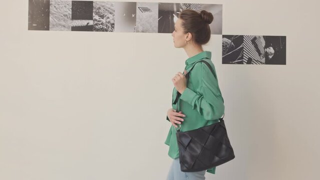 Medium slowmo portrait of young elegant Caucasian woman visiting modern photo gallery posing for camera standing against minimalist wall with black and white photos on it