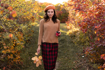 Autumn mood of happy smiling woman holding in her hands yellow leaf and coffee over orange-red foliage background