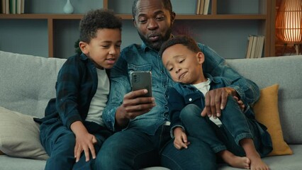 Happy African American family adult father with cute school kids children boys sons relax on sofa using funny smartphone apps talking use technology together looking at mobile phone play game at home - Powered by Adobe
