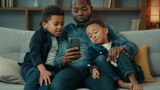 Happy African American family adult father with cute school kids children boys sons relax on sofa using funny smartphone apps talking use technology together looking at mobile phone play game at home