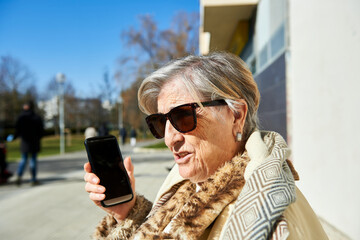 Fototapeta premium an eighty-year-old woman sunbathes in sunglasses sitting on a bench in front of a concrete wall while talking on the cellular phone