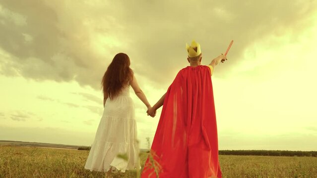 mother son walk through green field sunset. child teenager plays superhero nature. hold woman hand. teamwork. happy family. fancy dress red cloak, crown, sword. woman mother with boy son travels park