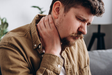 Close up of unhappy caucasian male holding hand on neck, suffering from neck pain, incorrect posture, joint inflammation. Unhealthy sad bearded man sitting on couch needs medicine chiropractor help