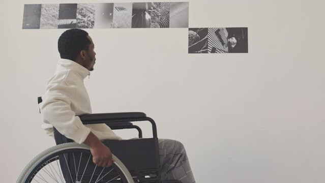 Slowmo of young African American man in wheelchair looking at pictures on walls in minimalist photo gallery