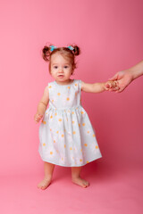 a baby girl stands on a pink background holding her mother's hand