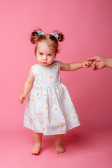 a baby girl stands on a pink background holding her mother's hand