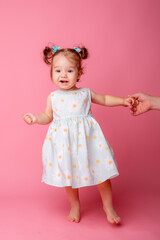 a baby girl stands on a pink background holding her mother's hand