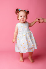 a baby girl stands on a pink background holding her mother's hand