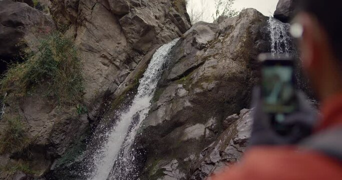 Slow motion of a hiker using his phone to take pictures of a waterfall flowing between rocky formations.