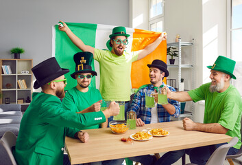 Group of happy, excited young and mature men friends with the Irish flag drinking green beer and having fun at a Saint Patrick's Day party at home. Celebration concept