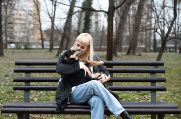 Young beautiful hipster woman sitting on a bench in the park with her adopted dog that saved his...