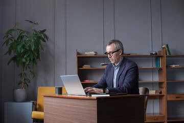 Mature pensive businessman with grey hair in glasses and suit is working on laptop in office