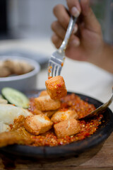 Close up sambal gami tahu tempe  or tempeh tofu with sambal cooked in earthenware plate against wooden background. Served on wooden table with chili, spoon and fork as decoration