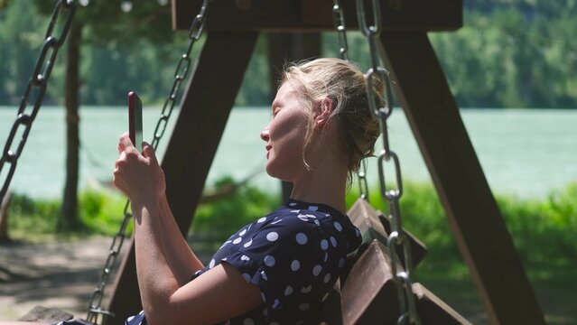 A young woman is sitting on a swing outdoors on a sunny summer day. Blonde woman holding a smartphone in her hands and smiling