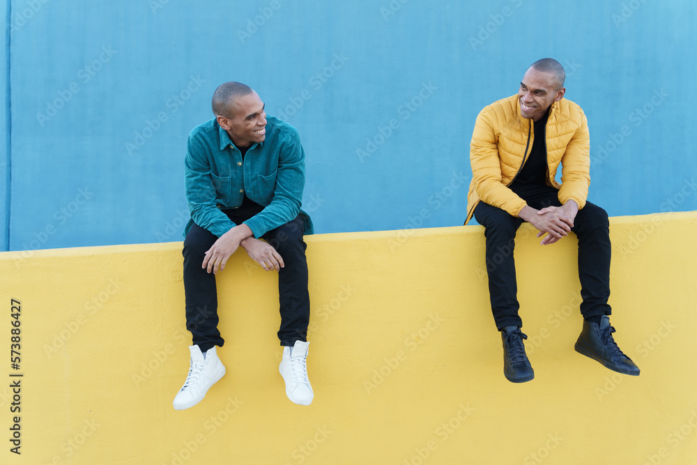 Wall mural smiling african american men brothers looking at each other while sitting on yellow concrete fence a