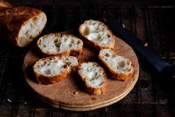 Sliced French baguette on a wooden board on a dark background. Bread with big holes. Preparing for bruschetta.