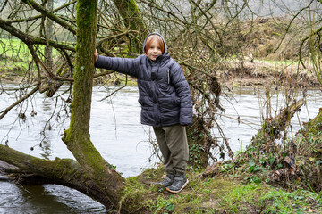 A young boy on a fallen tree near the river. A boy with long hair in a warm jacket is nine years...