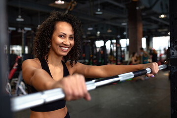 Beautiful young smiling african american woman looking at metal disc on barbell before hard bench press workout. Close-up.