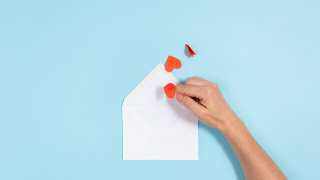Red paper hearts fly like butterflies to woman's hand and into an envelope. Blue background.