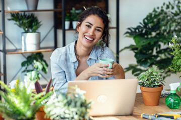 Shot of smiling woman drinking a cup of coffee while working with her laptop in greenhouse.