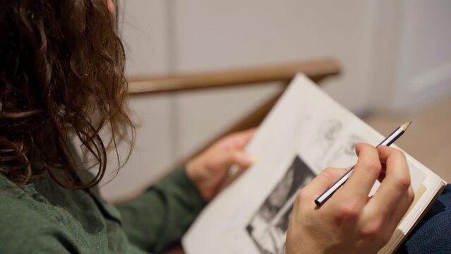 Profile shot of artist with long hair in green shirt drawing a face on sketch pad with pencil on sofa