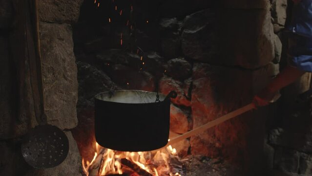 Woman raking buring logs in flames below cauldron in stone fireplace.